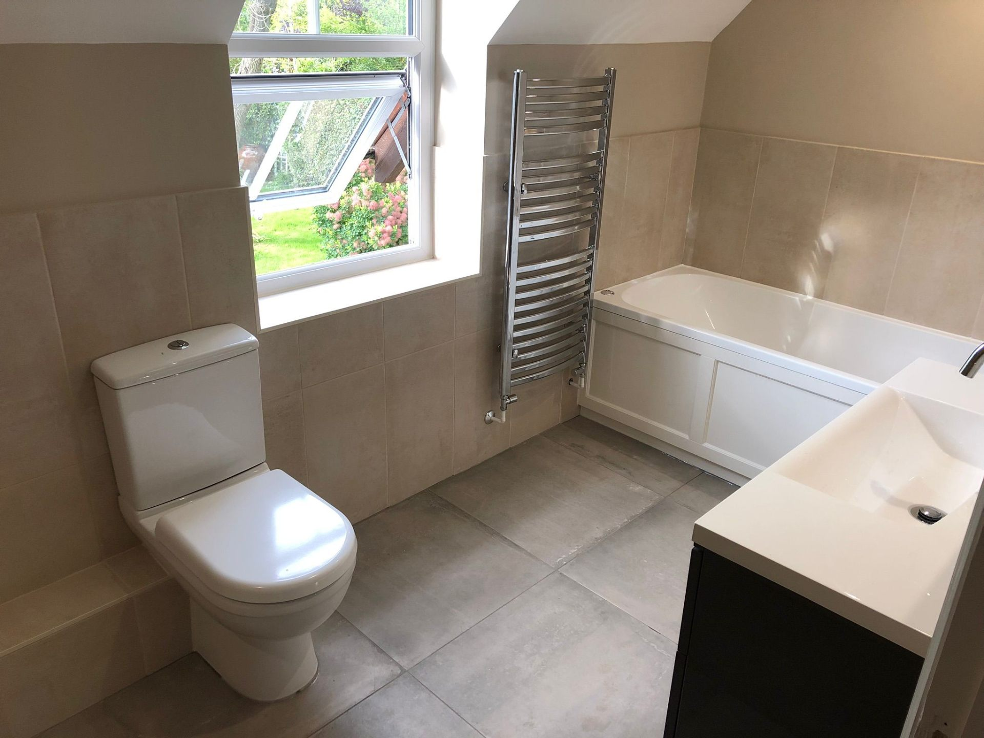 Modern bathroom with bath, chrome towel radiator, basin vanity and toilet fitted beneath a bright window